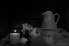 A monochrome photograph of a china wash jug and bowl with flowers and candle.